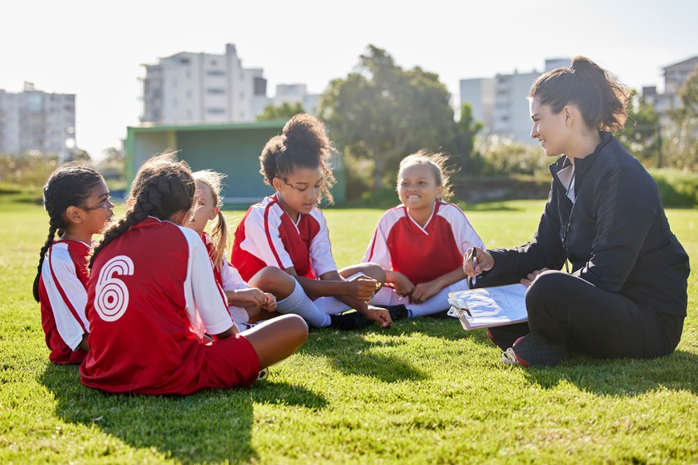 Female coach in a black jacket talking to young girls in red soccer uniforms on a field