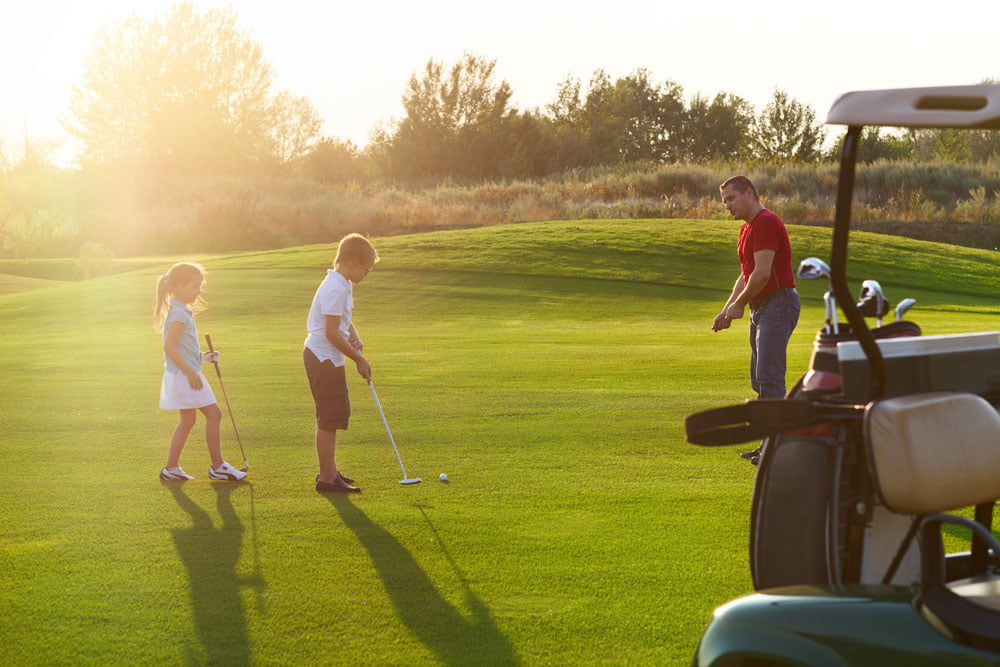 Young boy and girl learning golf with a male coach with golf cart in the foreground Young boy and girl learning golf with a male coach with golf cart in the foreground